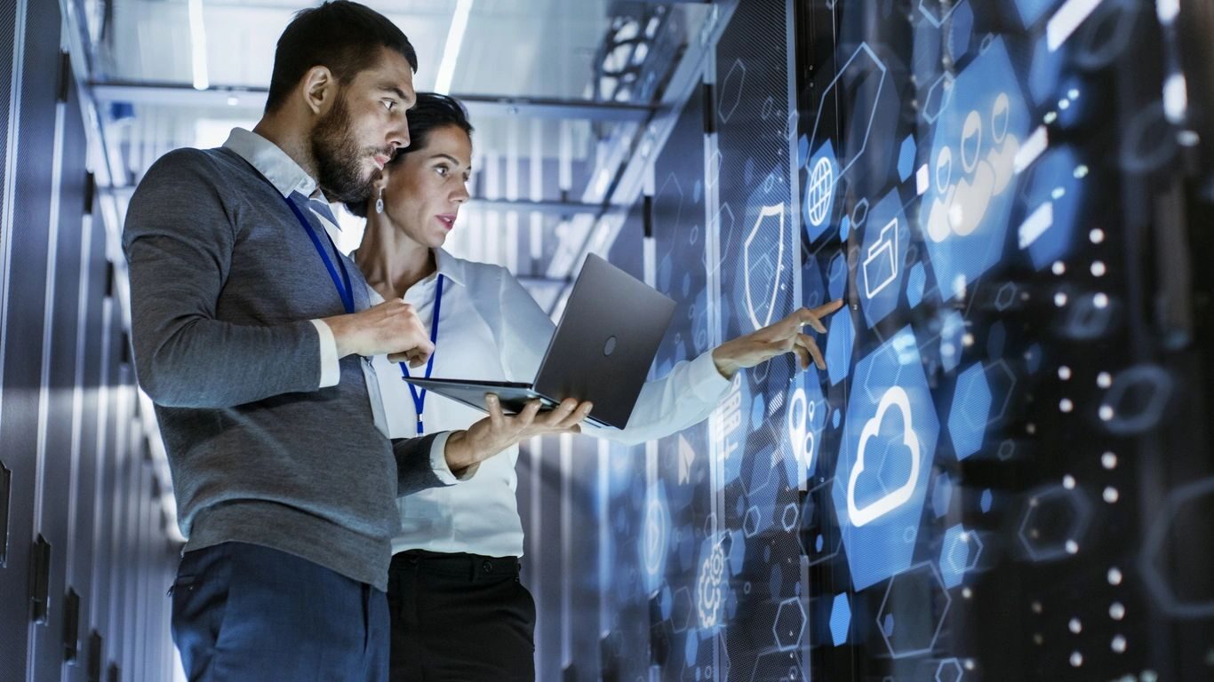 Two IT people looking at a big screen holding a laptop