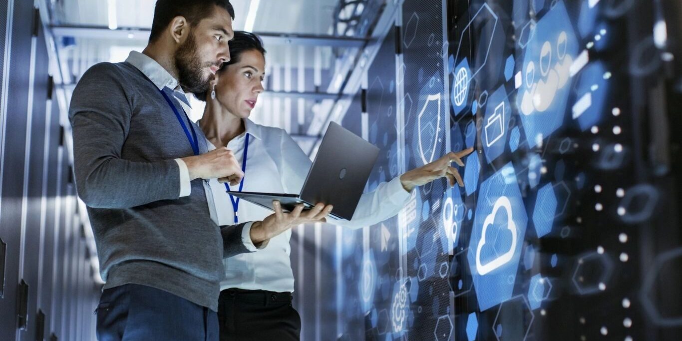 Two IT people looking at a big screen holding a laptop